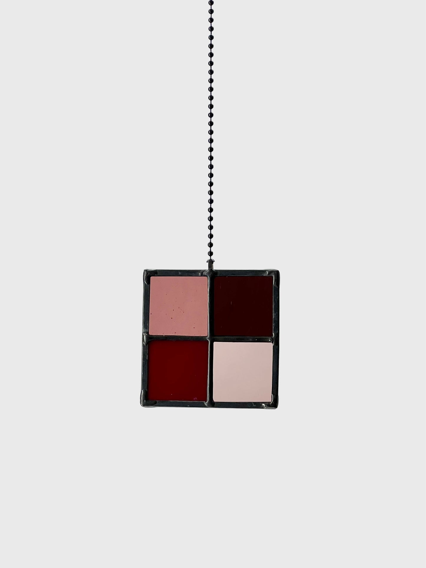 A square four-paned stained glass suncatcher in plum colors with a black frame, suspended by a black ball chain against a light gray background.